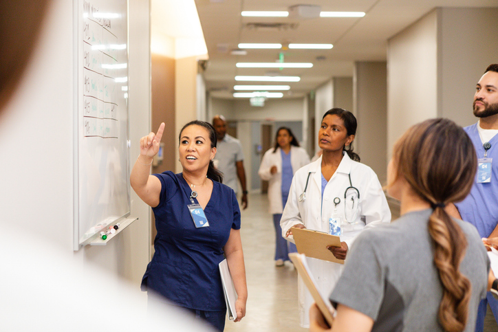 Young adult female nurse gestures towards the whiteboard while meeting with her team